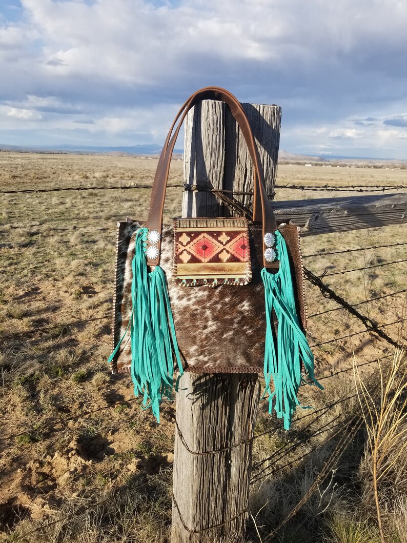 Puede incluir: Un bolso tote de cuero marr&oacute;n con flecos turquesa y un patr&oacute;n geom&eacute;trico colorido en la parte delantera. El bolso est&aacute; colgado en un poste de madera de una valla.