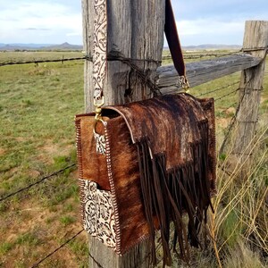 May include: A brown leather handbag with a fringe detail and a white floral patterned strap. The bag is hanging on a wooden fence post.