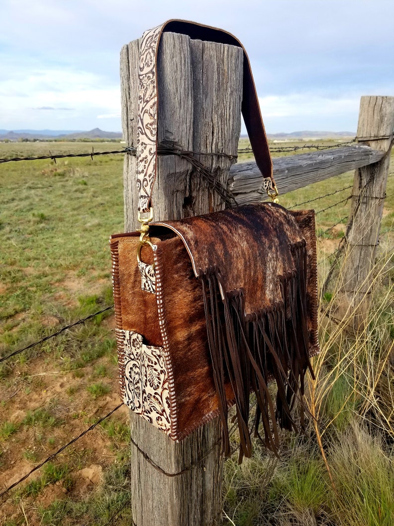 May include: A brown leather handbag with a fringe detail and a white floral patterned strap. The bag is hanging on a wooden fence post.