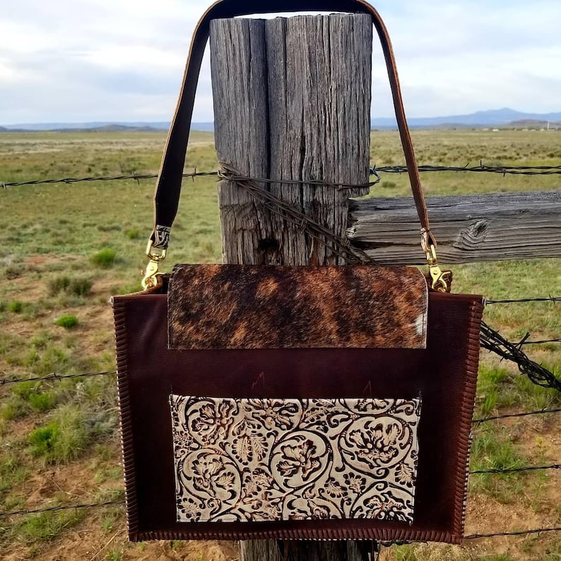 May include: A brown leather handbag with a cowhide flap and a decorative floral pattern on the front pocket. The bag has a long brown leather strap.