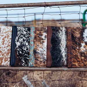 May include: Five cowhide journals with different patterns and colors, including brown, black, white, and turquoise, are displayed on a rusty wire fence.