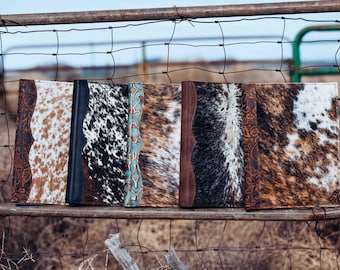 May include: Five rectangular journals featuring cowhide covers with varied patterns in brown, black, and white. Each journal includes a vertical spine accent with either embossed floral leather or a turquoise and copper geometric design.