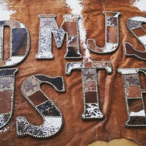 May include: A set of wooden letters covered in patchwork cowhide with a star design. The letters spell out the word "JUSTIN".