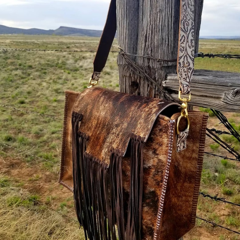 May include: A brown and white cowhide leather crossbody bag with a fringe detail and a brown leather strap. The bag is hanging on a wooden fence post.