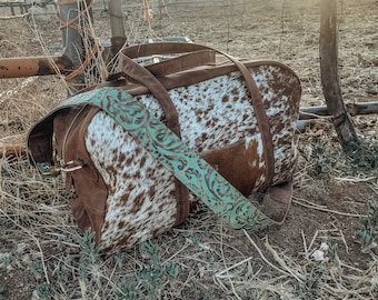 May include: A brown and white cowhide duffel bag with a turquoise floral patterned strap. The bag is sitting on a bed of dry grass.