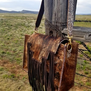 May include: A brown leather handbag with a fringe detail and a patterned strap. The bag is hanging on a wooden fence post.