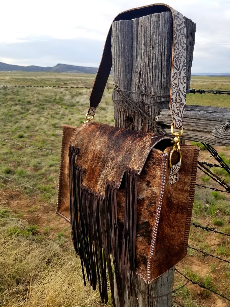 May include: A brown leather handbag with a fringe detail and a patterned strap. The bag is hanging on a wooden fence post.