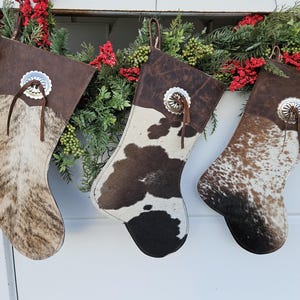May include: Three Christmas stockings with a Western theme. Each stocking features a different cowhide pattern in shades of brown, tan, and white. The top of each stocking is a dark brown leather with a silver concho and leather loop for hanging.