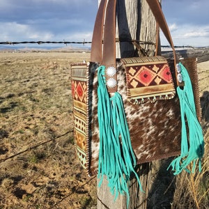 Puede incluir: Un bolso de mano de cuero marr&oacute;n con flecos turquesa y un patr&oacute;n de piel de vaca. El bolso est&aacute; colgado en un poste de madera con una valla de alambre de p&uacute;as en el fondo.