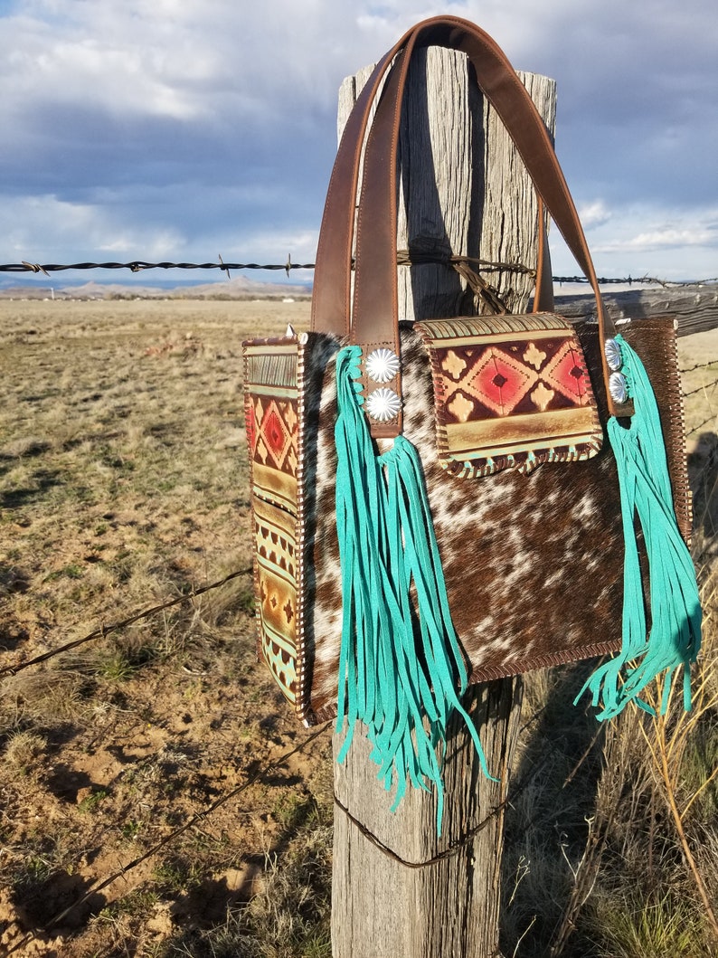 Puede incluir: Un bolso de mano de cuero marr&oacute;n con flecos turquesa y un patr&oacute;n de piel de vaca. El bolso est&aacute; colgado en un poste de madera con una valla de alambre de p&uacute;as en el fondo.