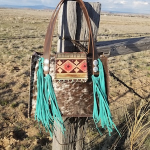 Puede incluir: Un bolso tote de piel de vaca marr&oacute;n y blanco con flecos turquesa y un bolsillo con estampado geom&eacute;trico. El bolso est&aacute; colgado de un poste de madera.