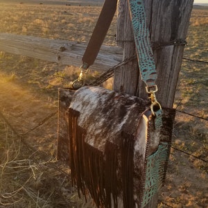 May include: A brown leather crossbody bag with a fringe detail. The bag is made of cowhide and has a turquoise leather strap with a snakeskin pattern.