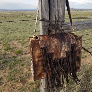 May include: A brown and white cowhide handbag with a long brown leather strap and fringe detail. The bag is hanging on a wooden fence post.