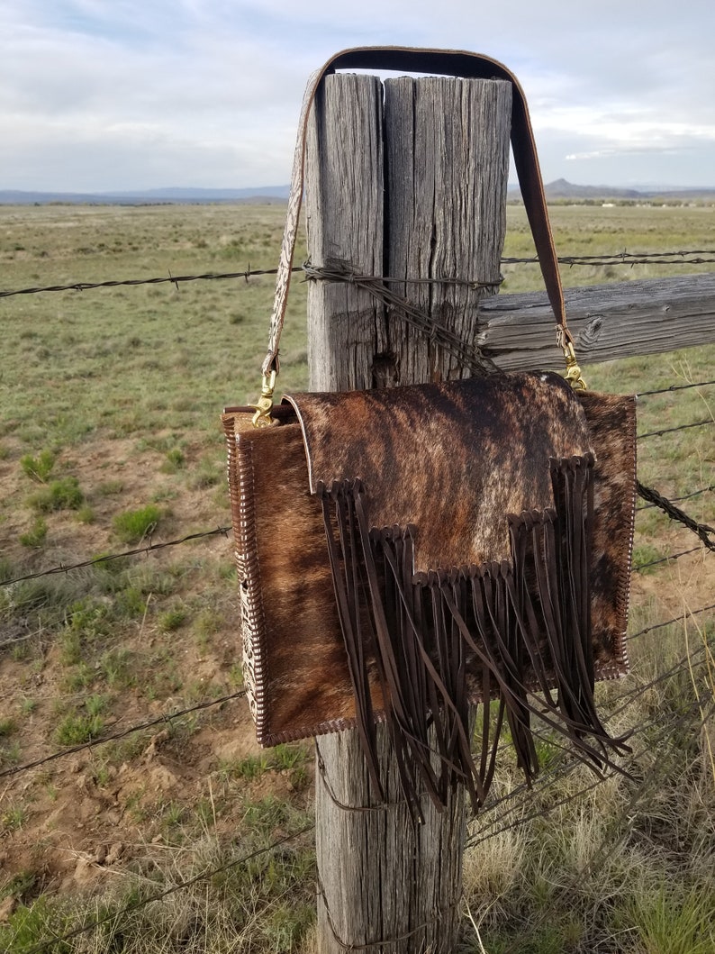 May include: A brown and white cowhide handbag with a long brown leather strap and fringe detail. The bag is hanging on a wooden fence post.