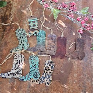 May include: A collection of ten leather boot ornaments with various patterns and colors, including turquoise, brown, and leopard print. The ornaments are hanging from twine strings against a rustic brown background.