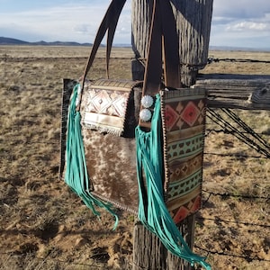 Puede incluir: Un bolso tote de cuero marr&oacute;n con flecos turquesa y un patr&oacute;n de piel de vaca. El bolso tiene un dise&ntilde;o geom&eacute;trico en la parte delantera y una hebilla plateada.