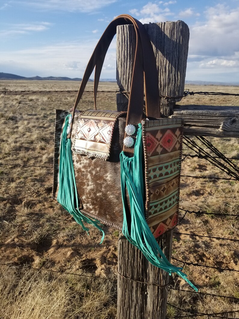 Puede incluir: Un bolso tote de cuero marr&oacute;n con flecos turquesa y un patr&oacute;n de piel de vaca. El bolso tiene un dise&ntilde;o geom&eacute;trico en la parte delantera y una hebilla plateada.