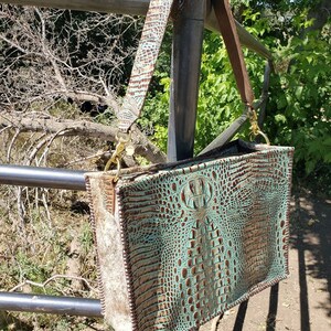 K&ouml;nnte beinhalten: Eine t&uuml;rkisfarbene und braune Ledertasche mit Kroko-Muster. Die Tasche hat einen langen braunen Lederriemen und goldene Beschl&auml;ge.