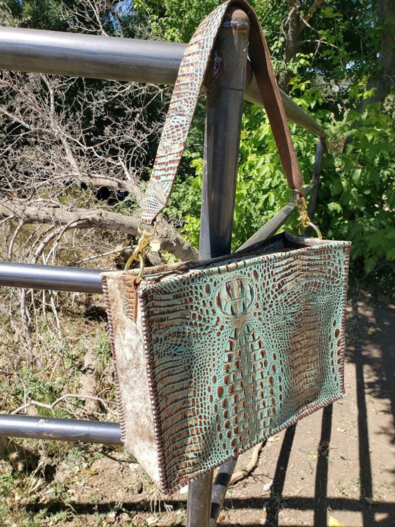K&ouml;nnte beinhalten: Eine t&uuml;rkisfarbene und braune Ledertasche mit Kroko-Muster. Die Tasche hat einen langen braunen Lederriemen und goldene Beschl&auml;ge.