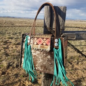 Puede incluir: Un bolso tote de cuero marr&oacute;n con flecos turquesa y un patr&oacute;n de piel de vaca blanca y marr&oacute;n. El bolso tiene una correa de cuero marr&oacute;n y una hebilla plateada.