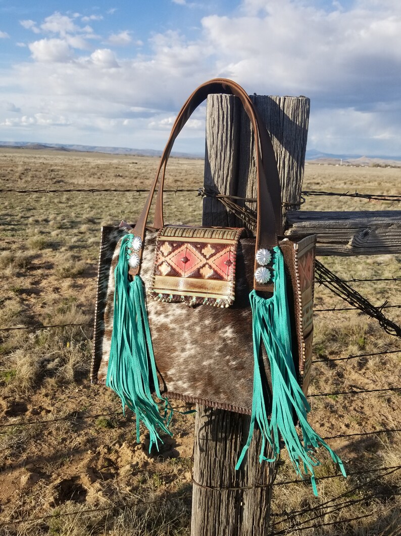 Puede incluir: Un bolso tote de cuero marr&oacute;n con flecos turquesa y un patr&oacute;n de piel de vaca blanca y marr&oacute;n. El bolso tiene una correa de cuero marr&oacute;n y una hebilla plateada.