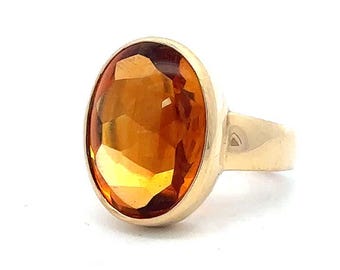May include: A gold ring featuring a large, oval-cut, amber-colored gemstone. The gemstone is faceted, catching the light and displaying a range of warm tones. The ring's band and setting are a polished gold color, creating a classic and elegant look.