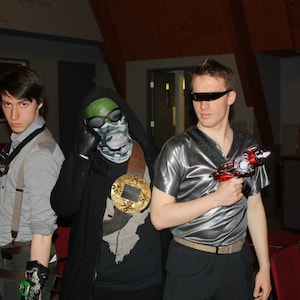 May include: Three people dressed in costumes, two men and one woman, are holding toy guns. The man on the left is wearing a grey shirt and a black vest. The man in the middle is wearing a black jacket and a green mask. The woman on the right is wearing a black shirt and a black jacket. The background is a dark room with a wooden ceiling.