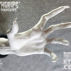 May include: White plaster hand with long, sharp fingernails, mounted on a black circular base. The hand is reaching out with fingers extended. The text "DEATHGRIPS™ WALL HANGERS" is visible at the top of the image. The text "DEATHGRIPS ETSY .COM" is visible at the bottom of the image.