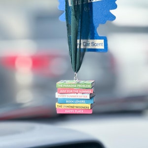 May include: A car air freshener with a blue pine tree design and the text "Trees" and "Car Scent". The air freshener is hanging from a rearview mirror and has a stack of miniature books attached to it. The books have titles such as "The Paradise Problem", "Just for the Summer", "Every Summer After", "Book Lovers", "The Unhoneymooners", and "Happy Place".