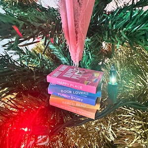 Op de afbeelding: Een stapel van vijf miniatuurboeken met kleurrijke omslagen, hangend aan een roze lint aan een kerstboom. De titels van de boeken zijn "Happy Place", "Book Lovers", "Funny Story", "Beach Read" en "People We Meet on Vacation".