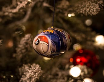 Könnte beinhalten: Ein Weihnachtsbaumschmuck in Form eines Pokéballs, mit einem blauen und weißen Design und einer roten und weißen Pokéball-Grafik. Der Schmuck hängt an einem Weihnachtsbaum.