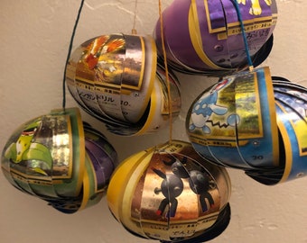 Könnte beinhalten: Eine Sammlung von sechs bunten, handgefertigten Ornamenten aus Pokémon-Sammelkarten. Jedes Ornament zeigt ein anderes Pokémon-Charakter, darunter ein grünes und gelbes Pokémon mit einer langen Zunge, ein rotes und gelbes Pokémon mit Flügeln, ein blaues und weißes Pokémon mit einem Schwanz, ein violettes und gelbes Pokémon mit einem Schwanz, ein schwarzes und weißes Pokémon mit einem Schwanz und ein goldenes und schwarzes Pokémon mit einem Schwanz. Die Ornamente hängen an Schnüren.