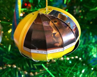 Könnte beinhalten: Ein gelber Weihnachtsbaumschmuck aus einer Pokemon-Sammelkarte mit einem Pikachu, das eine Tasse hält. Die Kartennummer ist 025 und der Name des Pokemon ist "Pikachu".