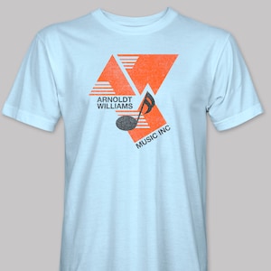 May include: Light blue t-shirt with an orange and black graphic design. The graphic features a musical note and the text "Arnoldt Williams Music Inc."
