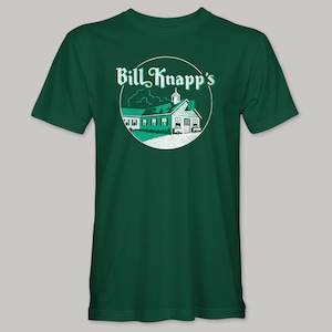 May include: A dark green t-shirt featuring a circular graphic with the text "Bill Knapp's". The graphic depicts a white building with a green roof and accents, set against a backdrop of clouds and a steeple.