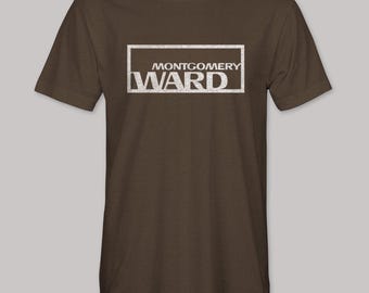 May include: Brown t-shirt featuring the text "MONTGOMERY WARD" in a white rectangular frame. The text has a distressed, vintage appearance. The shirt has short sleeves and a crew neck.