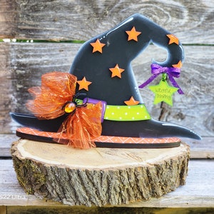 Puede incluir: Un sombrero de bruja negro con detalles en naranja y verde, un lazo morado y una etiqueta en forma de estrella que dice "Witcheful Thinking". El sombrero está sentado sobre una rodaja de tronco de madera.