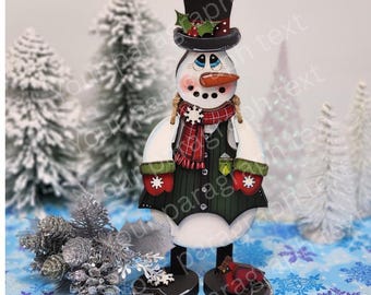 Op de afbeelding: Houten sneeuwman decoratie met een zwarte hoge hoed, wortelneus en een rood-zwarte geruite sjaal. De sneeuwman draagt een groen vest en rode wanten met witte sneeuwvlokontwerpen. De basis is zwart met rode accenten.