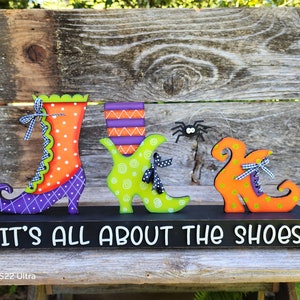 Puede incluir: Tres zapatos de bruja de madera pintados en naranja, verde y morado, con lunares negros y una araña colgando sobre ellos. Los zapatos están sobre una base de madera negra con el texto "It's All About The Shoes".