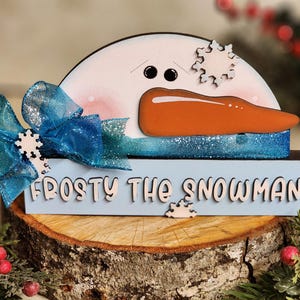May include: A wooden snowman figurine with a blue and white bow, a carrot nose, and black eyes. The snowman is sitting on a wooden log with the words "Frosty the Snowman" written on a blue sign.