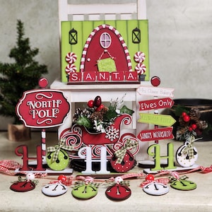 以下が含まれることがあります： サンタクロースの赤い白のそりと、北極の看板、そして「ミセス・クロースのベーカリー、エルフのみ、トナカイに乗る、サンタのワークショップ」と書かれた看板があるクリスマスの村のシーン。赤、白、緑のアクセントがついたジングルベルもいくつかあります。