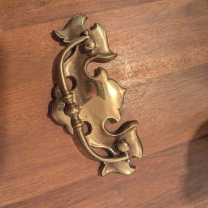 May include: A polished brass drawer pull featuring a decorative, ornate backplate with scalloped edges and a curved, swinging bail handle. The hardware is finished with a metallic gold tone and includes two circular mounting points for attachment.