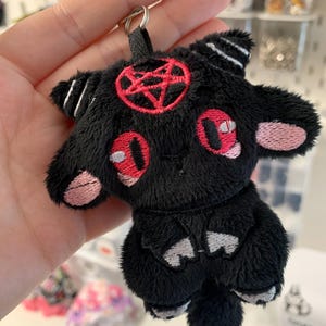 May include: Black plush keychain in the shape of a cartoonish goat with a red pentagram on its head. The goat has red eyes and pink accents on its ears and snout. The keychain has a black loop for attaching to keys or a bag.