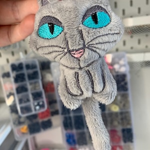 May include: A gray plush cat keychain with bright turquoise eyes and a pink nose. The cat has embroidered details and a fluffy texture. The keychain includes a metal ring for attaching to keys or bags.