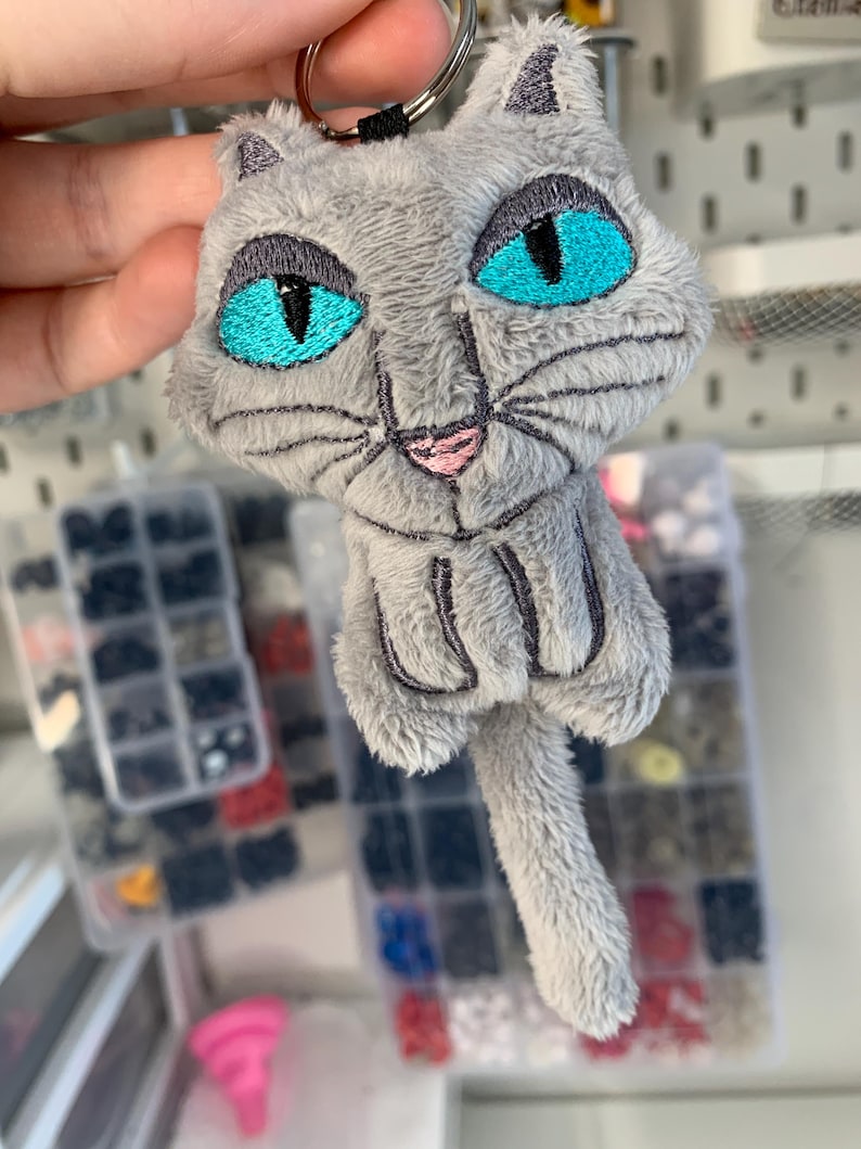 May include: A gray plush cat keychain with bright turquoise eyes and a pink nose. The cat has embroidered details and a fluffy texture. The keychain includes a metal ring for attaching to keys or bags.