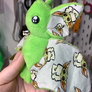 May include: A plush green bat-like creature with large black eyes and patterned wings. The wings feature a repeating image of a character with large ears and a neutral expression. The plush toy is soft and cuddly.