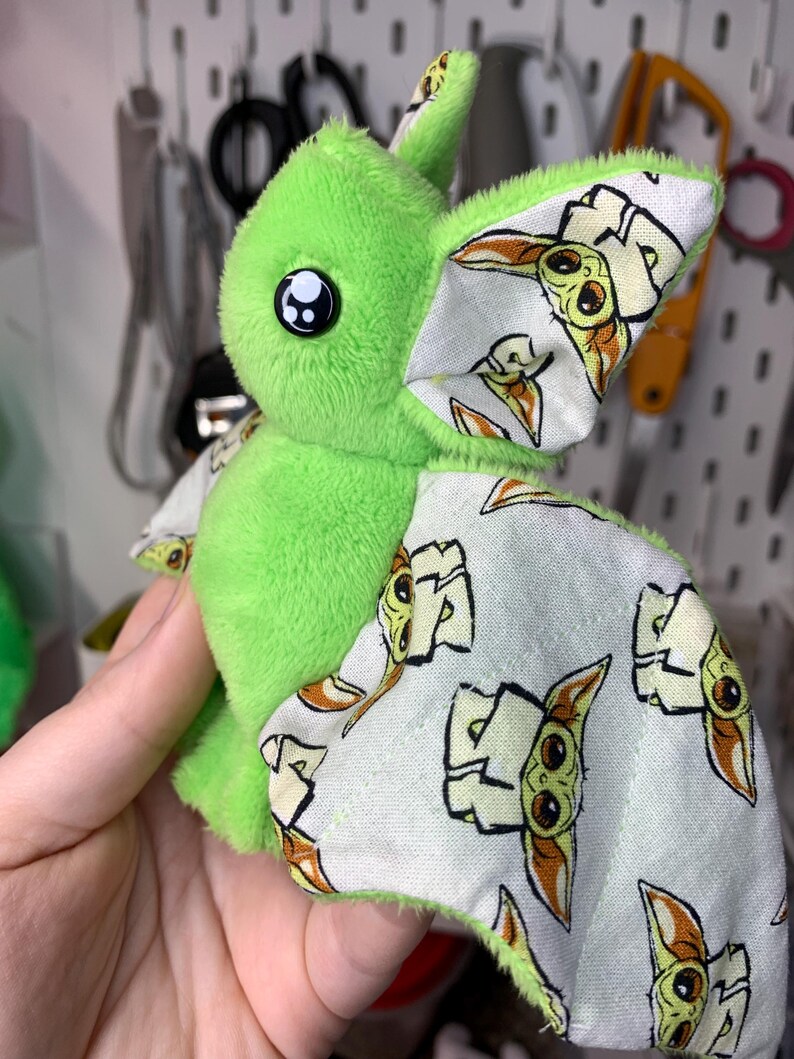 May include: A plush green bat-like creature with large black eyes and patterned wings. The wings feature a repeating image of a character with large ears and a neutral expression. The plush toy is soft and cuddly.
