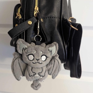May include: A grey plush bat charm with embroidered details, attached to a black backpack. The bat has large ears, wings, and a smiling face. The backpack has gold zips and a top handle.