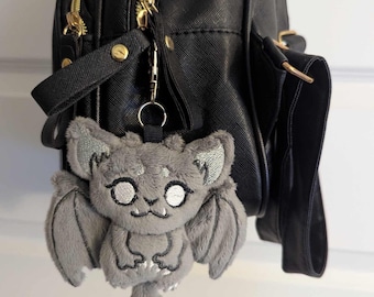 May include: A gray plush bat charm with embroidered details, attached to a black backpack. The bat has large ears, wings, and a smiling face. The backpack has gold zippers and a top handle.