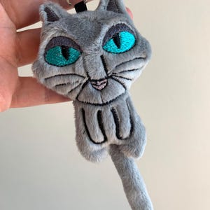 May include: A gray plush cat keychain with embroidered details. The cat has large teal eyes, a pink nose, and black whiskers. The keychain includes a silver metal ring and a black fabric loop.